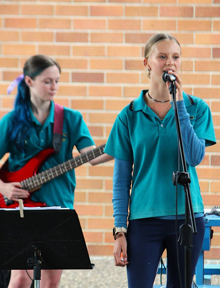 A female student singing into a microphone on stage while another student plays electric bass behind her.